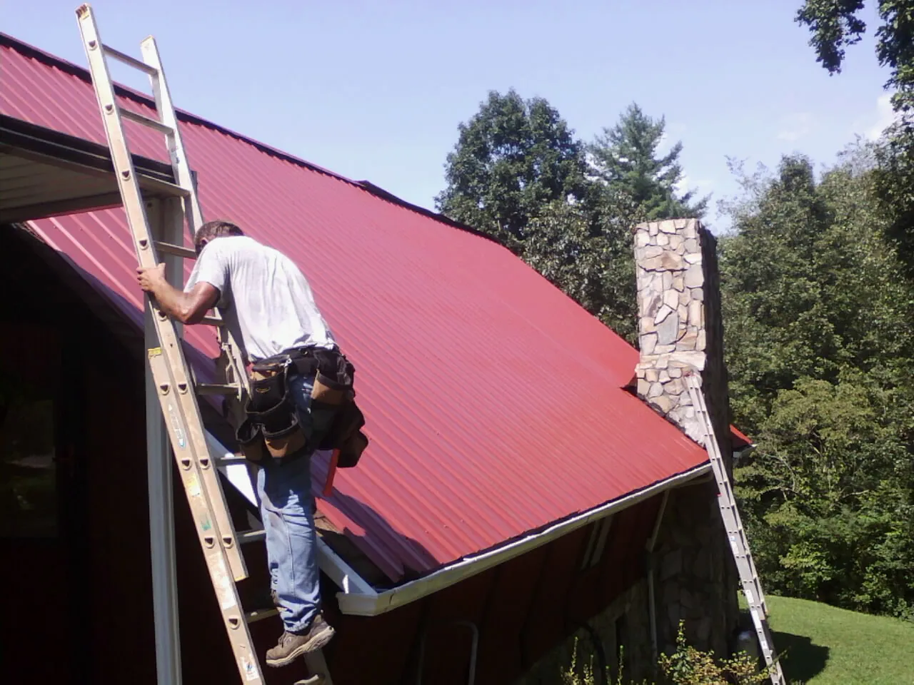 Roofer on a ladder beside a red metal roof and stone chimney — Soffit & Fascia Repair by Roofing Brothers in Chancellor
